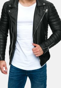 Zwarte leren motorjack met gequiltte schouders, zilveren ritsen en een wit T-shirt eronder, gecombineerd met blauwe jeans.