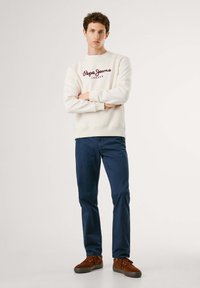 Sudadera crema con logo burdeos, pantalones azul marino y zapatos marrones. El modelo está de pie con los brazos cruzados en un fondo neutro.