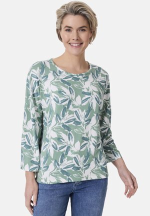 Langarm-Top in Grün mit weißem Blumenmuster, Rundhalsausschnitt und lockerer Passform. Hergestellt aus weichem, leichtem Stoff. Kombiniert mit blauen Jeans.