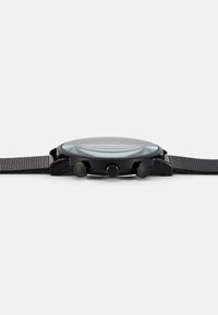 Montre noire avec un boîtier rond en quartz, trois boutons, un bracelet texturé et un cadran en verre clair. Design minimaliste avec un profil élancé.