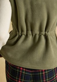 Tuva kāda persona, kura valkā olīvgarenas krāsas vilnas vesti pār krēmkrāsas džemperi un tumšas pārbaudītas bikses.