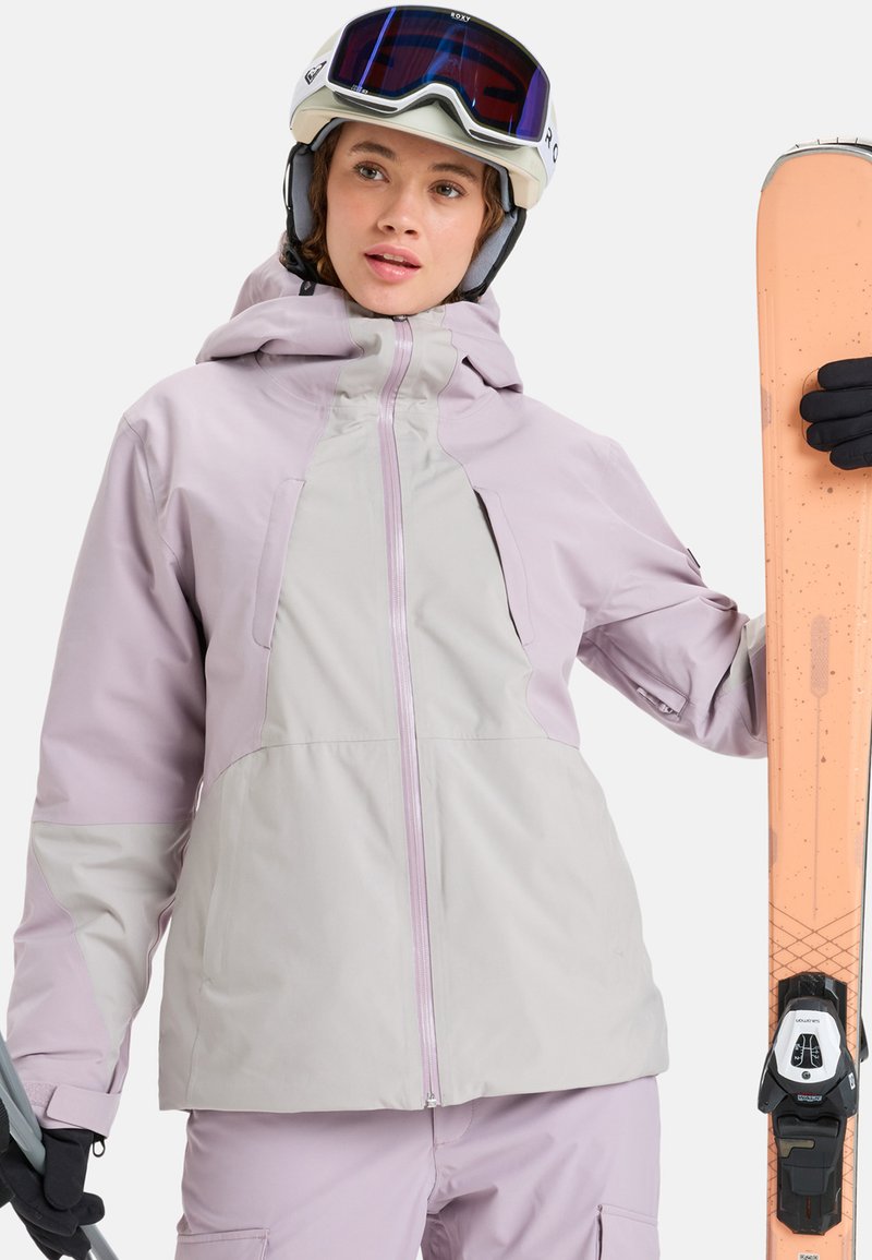 Veste de ski en lavande douce et gris clair, avec une fermeture éclair à l'avant, une capuche et de multiples poches. Équipement de ski en main.