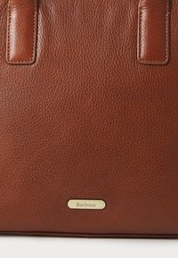 Bruine leren handtas met een gestructureerd oppervlak, voorzien van een gouden label met de tekst "Barbour" en stevige, gestikte handvatten.
