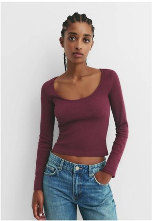 Mladá žena so zapletenými vlasmi, oblečená v priliehavé bordové tričko s dlhým rukávom a modré džínsy, stojaca na neutrálnej pozadí.
