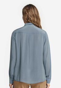Blusa a maniche lunghe azzurro chiaro con una texture liscia, con polsini abbottonati e orlo curvo. Presenta un yoke posteriore e una discreta cucitura decorativa.