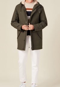 Parka olive verte avec capuche en fourrure, fermetures à bouton et à zip, poches latérales, portée sur un pull rayé et un pantalon blanc.