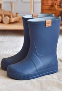 Een paar blauwe rubberen regenlaarzen met een lichtgrijze voering, geplaatst op een beige oppervlak met op de achtergrond een houten speelgoed en een mand.