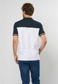 Polo skjorte med en marineblå top og hvid bund. Har korte ærmer med røde og blå striber. Materialet ser let og struktureret ud.