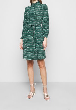Femme portant une robe verte à manches longues à motifs avec une ceinture à la taille et des sandales blanches à talons avec brides, debout devant un fond uni.
