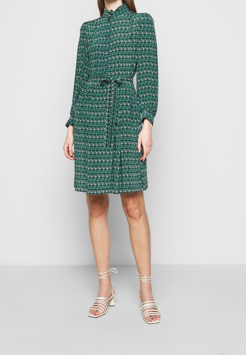 Femme portant une robe verte à manches longues à motifs avec une ceinture à la taille et des sandales blanches à talons avec brides, debout devant un fond uni.