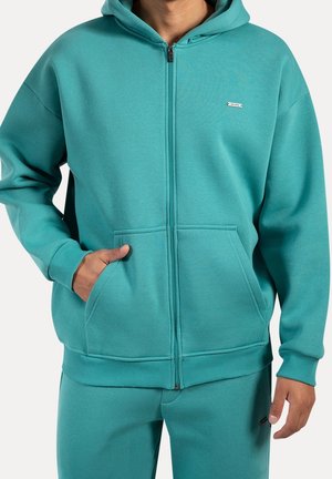 Person trägt einen petrolfarbenen Kapuzenpullover mit Reißverschluss und Fronttaschen sowie passende petrolfarbene Hose, Hand in der Tasche, vor weißem Hintergrund.