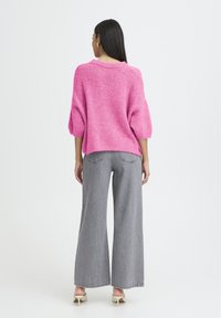 Femme aux longs cheveux bruns portant un pull rose texturé, un jean gris à jambe large et des chaussures à talons blancs, se tenant devant un fond blanc uni.