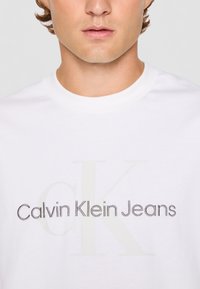 T-shirt bianca in cotone con la scritta "Calvin Klein Jeans" in tonalità di grigio e nero, con un logo "CK" prominente sullo sfondo.