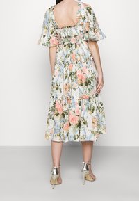 Vestido midi floral con mangas abullonadas en blanco, que presenta flores rosas y azules, un respaldo fruncido y un borde de volantes en capas. Sandalias de tacón alto plateadas.