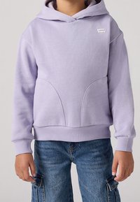 Sudadera lavanda con bolsillo canguro, puños y dobladillo acanalados, que presenta un pequeño logo de Levi's en el pecho. Combinada con pantalones cargo de mezclilla azul.