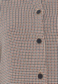 Vävt tyg med ett hundtandsmönster i marinblått, krämfärgat och rostfärgat, med cirkelrunda svarta knappar längs framsidan.