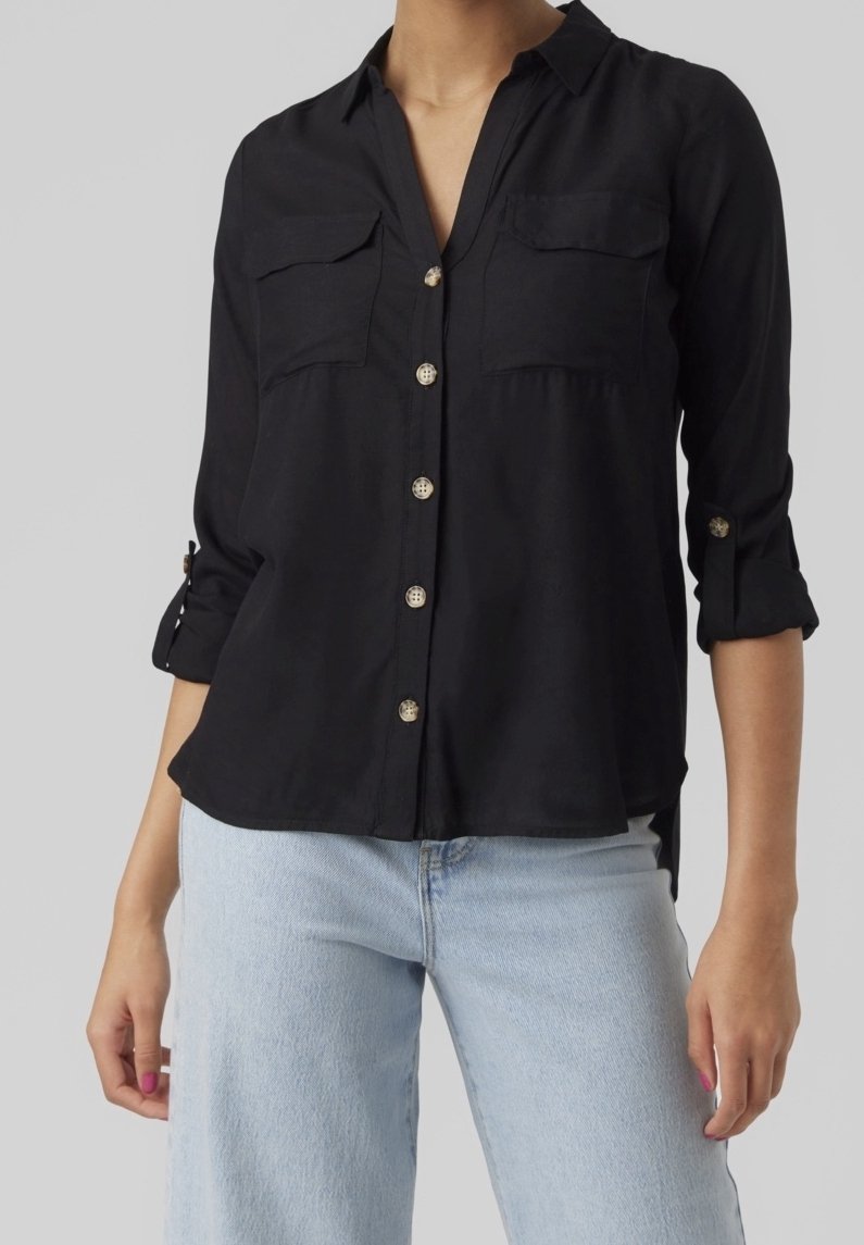 Femme portant une chemise noire à boutons avec manches retroussées et un jean bleu clair, montrée des épaules jusqu'aux hanches sur fond gris.