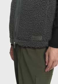 Gilet en polaire gris avec un tissu texturé, fermeture éclair à l'avant et patch logo. Associé à un pantalon vert foncé avec des poches.