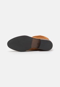 Botte avec un dessus en daim marron, bout rond, semelle en caoutchouc noir texturé et coutures détaillées sur les côtés.