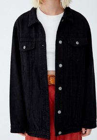 Schwarze Denimjacke mit durchgehender Knopfleiste, zwei Brusttaschen und silberner Hardware. Verfügt über einen lockeren Schnitt und lange Ärmel.