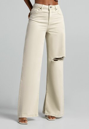 Beżowe dżinsy o szerokich nogawkach z wysokim stanem, dużymi przednimi kieszeniami i przetarciem na kolanie. Wykonane z denimu o gładkiej fakturze.