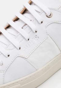 Baskets blanches en cuir avec des détails texturés, présentant des lacets plats blancs, un logo argenté et une semelle en caoutchouc beige clair.