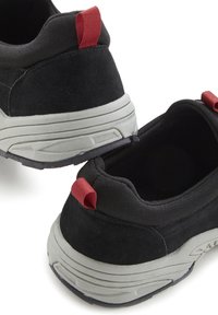 Schwarze Sportschuhe mit Wildledertextur, grauer Sohle und rotem Zugschlaufe an der Ferse. Abgerundetes Design, gepolsterter Schaft und minimales Profil.