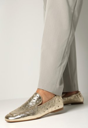 Metallic gouden loafers met een gladde textuur, voorzien van ronde uitsparingen en een ronde teenvorm. Gedragen met lichtgrijze broek.