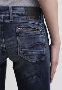 Jeans de mezclilla azul oscuro con un ajuste slim, que cuentan con bolsillos traseros, un pequeño bolsillo con cremallera y una etiqueta de cuero en la cinturilla. Tela texturizada con desgastes.