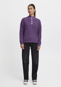 Paarse gebreide trui met een hoge halslijn en vier witte knopen langs de sluiting, gecombineerd met zwarte rechte broek en sneakers.