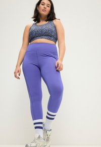 Paarse leggings met hoge taille, gecombineerd met een cropped zwart shirt met een blauw luipaardpatroon. Zilveren sneakers met witte en blauwe gestreepte sokken.
