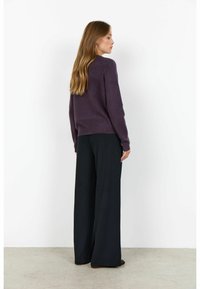 Femeie cu părul lung castaniu deschis, îmbrăcată cu un pulover violet închis și pantaloni largi negri, stând cu fața către un perete alb.