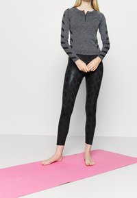 Kvinna stående barfota på en rosa yogamatta, iförd en grå långärmad topp med svarta ränder och glänsande svarta leggings med ett ormskinnsmönster.