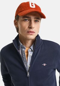 Giacca a zip blu navy con colletto, con un piccolo logo. Sotto, una camicia a quadri blu chiaro e una maglietta arancione. Cappello in velluto a coste rosso.