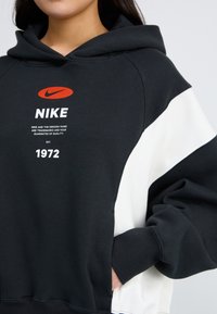Haut à capuche surdimensionné en noir et blanc avec une poche kangourou, logo Nike en orange et blanc bien en vue, poignets côtelés et tissu texturé.