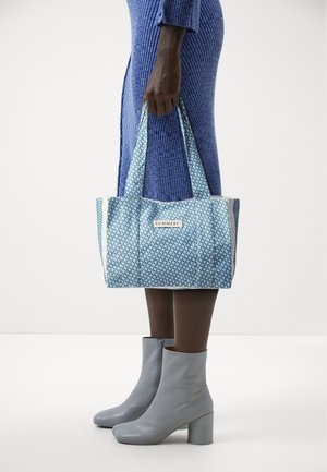 Personne en robe bleu côtelée tenant un sac fourre-tout estival bleu à motifs, portant des bottines gris clair à talons blocs.