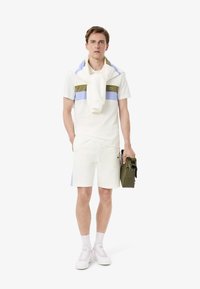 T-shirt polo blanc avec des rayures horizontales vert olive et bleu clair, associé à un short blanc et des baskets blanches. Tenant un sac vert.