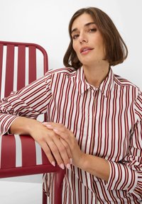 Striped shirt in red and white fabric, featuring a button-up design. A red metal chair with slatted backrest is in the background.