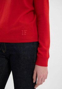 Pull rouge en tricot avec des manches longues, présentant un ourlet et des poignets côtelés. Subtil logo "IF" brodé près du bas. Porté avec un jean sombre.