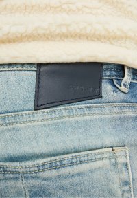 Vaqueros de mezclilla azul claro con un parche de cuero con la etiqueta "CHASING." Presentan detalles de costuras visibles y una capa texturizada de algodón encima.