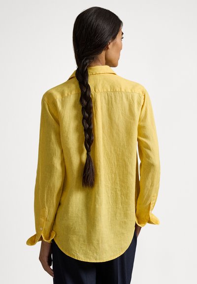 Sieviete ar gariem pītajiem matiem valkā vaļīgu dzeltenu lina kreklu un tumšas bikses, stāvot ar muguru pret balto fonu.