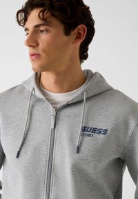 Sweat à capuche gris avec fermeture éclair et cordons, arborant un logo bleu "GUESS" et l'année "1981" brodés sur la poitrine. Tissu doux et texturé.