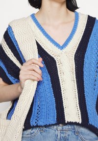 Pull en tricot oversize rayé bleu, marine et crème. Design en V avec des détails texturés et des bords côtelés, associé à un sac crème.