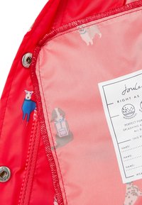 Den röda vattentäta jackans insida har en ljusrosa foder med illustrerade hundar i färgglada kläder, tillsammans med en omsorgsetikett som är tydligt synlig.