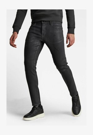 Uomo che indossa jeans slim-fit neri con leggeri effetti di usura, abbinati a sneaker nere con suole bianche, in piedi su sfondo chiaro.