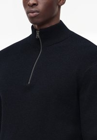 Pull noir à demi-fermeture éclair avec col côtelé porté par une personne, fermeture partielle, focus sur le haut du torse et le cou.
