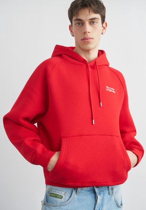 Mladić u svijetlo crvenoj majici s kapuljačom i prednjim džepom s natpisom „the new originals“, uparenom s svijetloplavim trapericama.