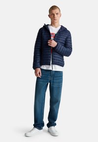 Mladý muž v námořnické modré péřové bundě přes grafické bílé tričko, modré džíny a bílé tenisky, stojící na bílém pozadí.