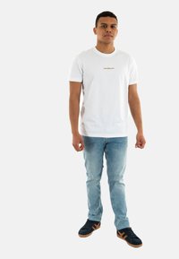 Camiseta de algodón blanca con un pequeño logo dorado, combinada con jeans de mezclilla azul claro y zapatillas negras con acentos marrones.