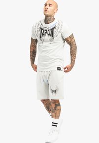 Weißes TAPOUT T-Shirt mit schwarzem geflügeltem Logo und blauer Schrift, kombiniert mit passenden Shorts. Mit Kordelzug und grafischen Akzenten auf beiden Teilen.